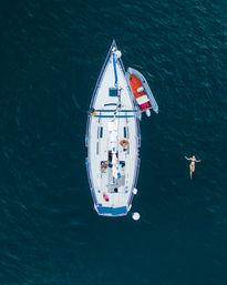 Aerial drone view of a white sailboat anchored in deep blue water with a small dinghy tied alongside and a person floating nearby.
