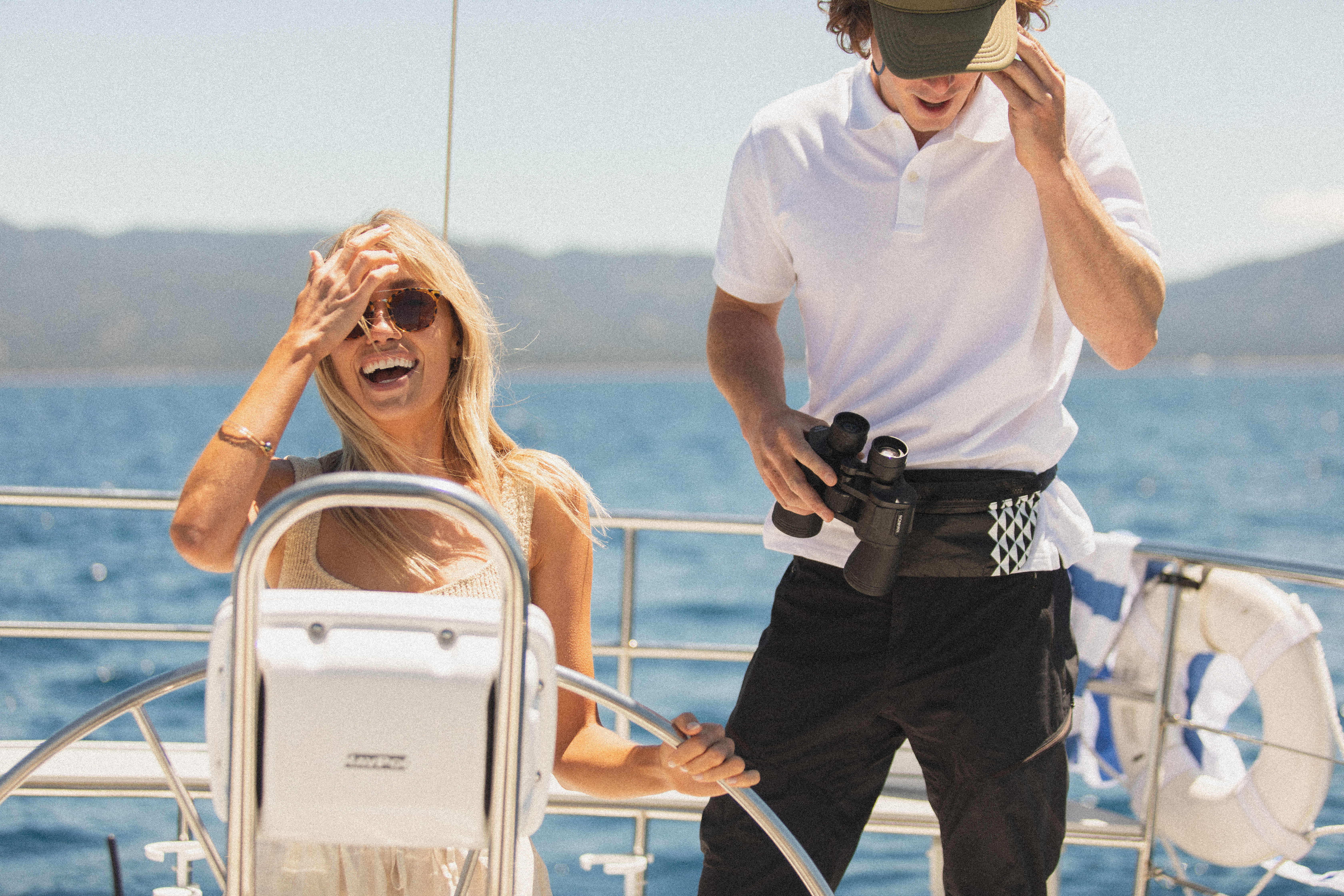 Smiling woman at a sailboat helm wearing sunglasses while a crew member in a polo holds binoculars on a sunny day over blue coastal waters.