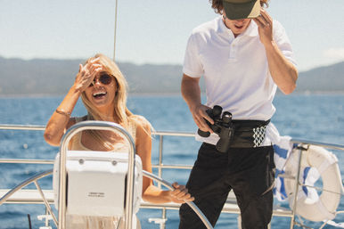 Smiling woman at a sailboat helm wearing sunglasses while a crew member in a polo holds binoculars on a sunny day over blue coastal waters.