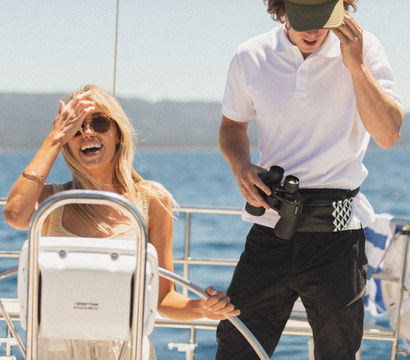 Smiling woman at a sailboat helm wearing sunglasses while a crew member in a polo holds binoculars on a sunny day over blue coastal waters.