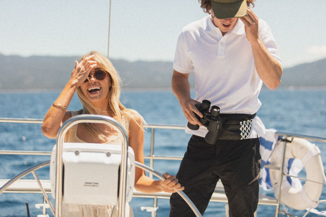 Smiling woman at a sailboat helm wearing sunglasses while a crew member in a polo holds binoculars on a sunny day over blue coastal waters.