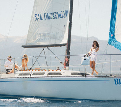 Three people enjoying a sail on an alpine lake: a woman in a white cover-up stands at the bow holding a striped tote while two others relax on the deck under tall white sails with sunlit mountains in the background.