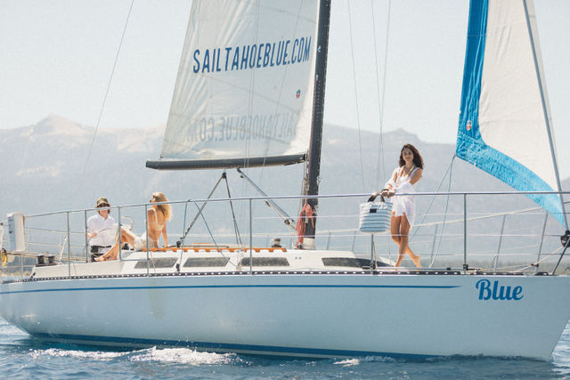 Three people enjoying a sail on an alpine lake: a woman in a white cover-up stands at the bow holding a striped tote while two others relax on the deck under tall white sails with sunlit mountains in the background.