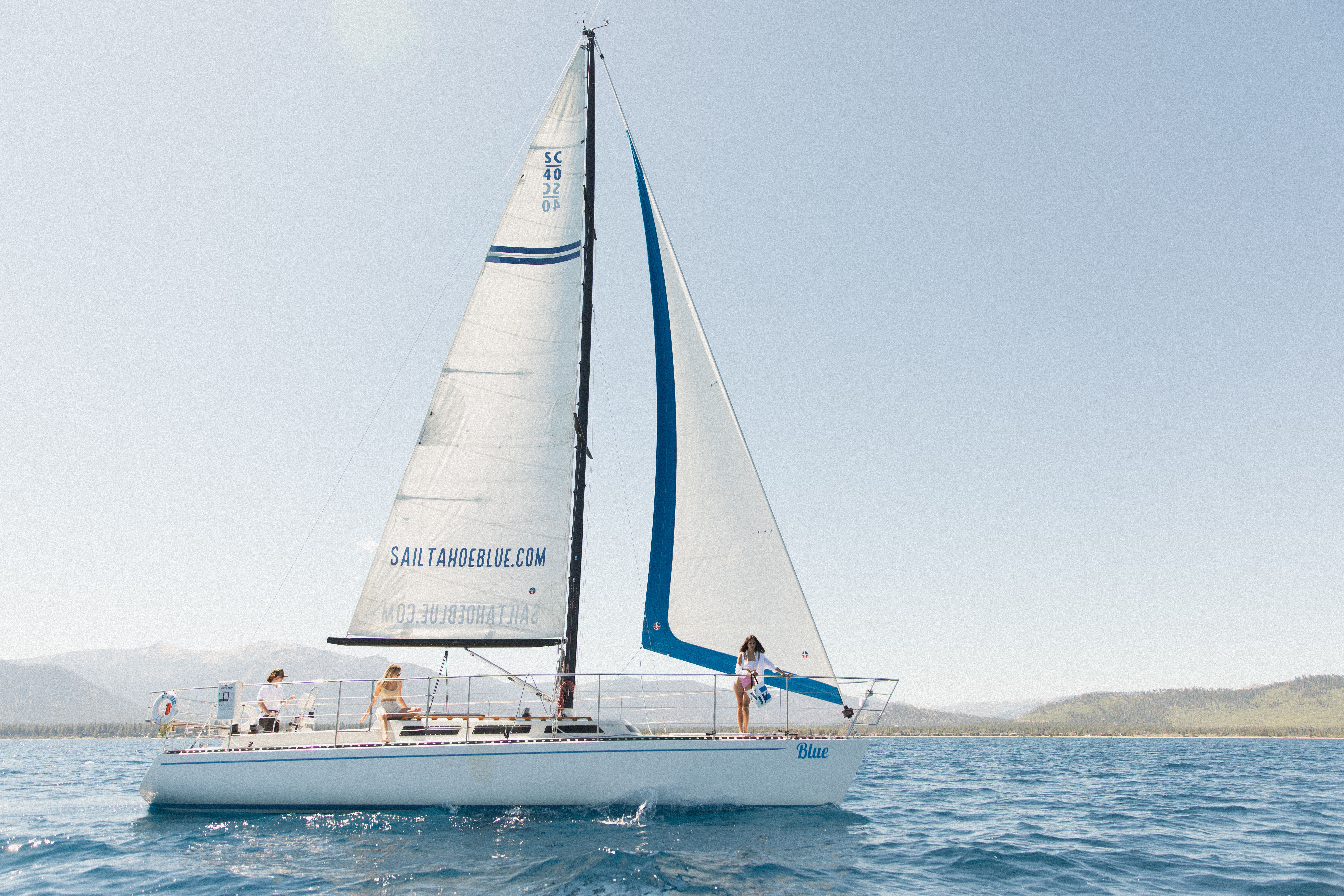 White sailboat with blue accents and three people on deck sailing on a sunny alpine lake with a tree-lined shore and distant mountains.