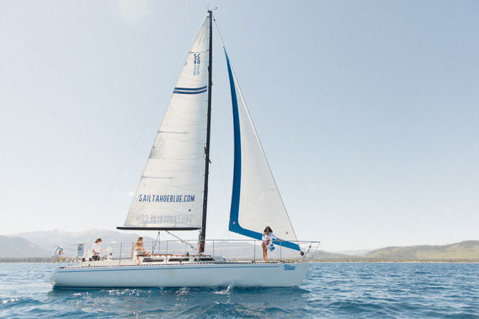 White sailboat with blue accents and three people on deck sailing on a sunny alpine lake with a tree-lined shore and distant mountains.