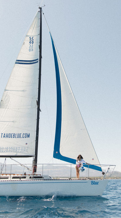 White sailboat with blue accents and three people on deck sailing on a sunny alpine lake with a tree-lined shore and distant mountains.