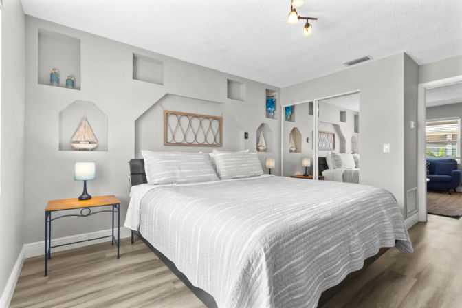 Bright modern master bedroom with king bed in gray striped bedding, built-in wall niches featuring nautical decor, mirrored sliding closet doors, wood-look flooring and a bedside table with blue lamp.