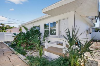 Cheerful white single-story coastal cottage with a small porch marked 5, lush tropical palms and greenery along a stone walkway under a bright blue sky.