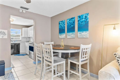Coastal-style dining nook in an open-plan apartment: white high-top table with four cushioned chairs, blue ocean triptych on beige wall, view into white kitchen with stainless appliances and tile floor.