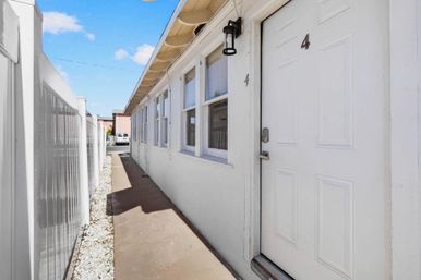 Sunlit narrow exterior walkway beside a white stucco apartment building with a row of windows, a white door marked 4, black wall lantern, and a tall white vinyl fence under a blue sky.