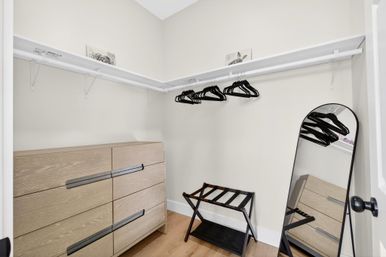 Minimal modern walk-in closet with light wood dresser, black hangers on a white rod, folding luggage rack and leaning full-length mirror on hardwood floor in neutral tones.
