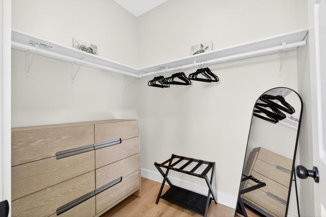 Minimal modern walk-in closet with light wood dresser, black hangers on a white rod, folding luggage rack and leaning full-length mirror on hardwood floor in neutral tones.
