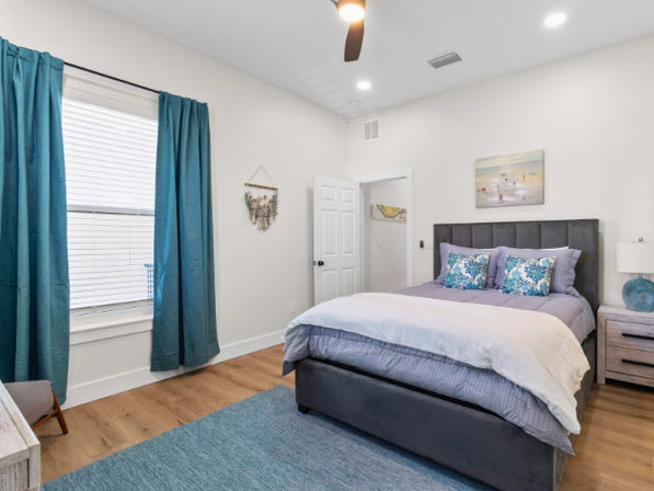Bright modern bedroom with gray upholstered bed, teal curtains and accent pillows, hardwood floors, teal area rug, nightstand, ceiling fan and beach-themed wall art.