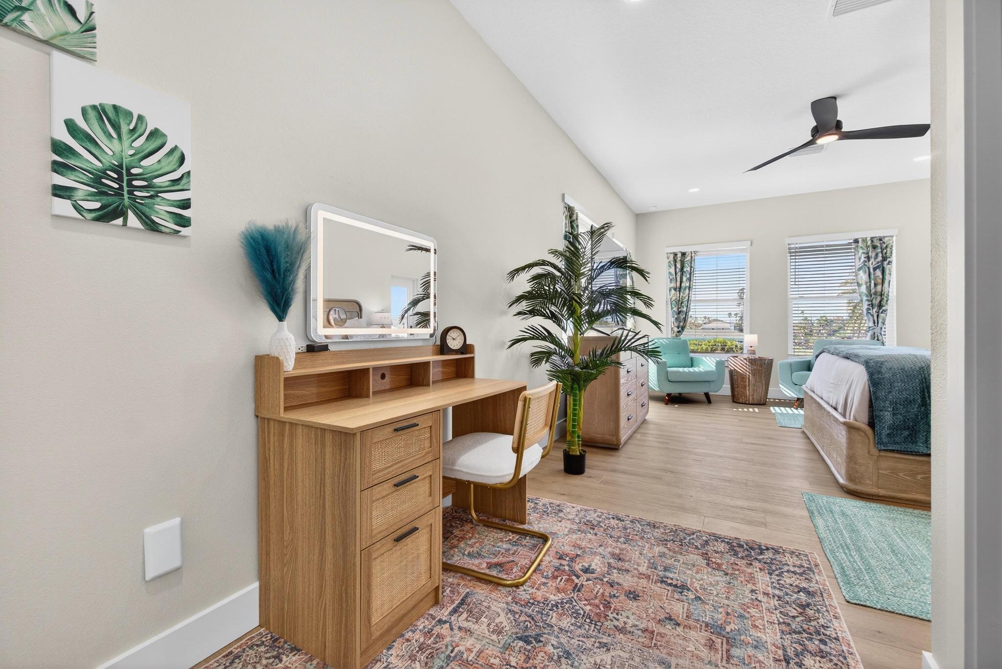 Sunlit coastal-style master bedroom with hardwood floors, wooden vanity desk with rattan drawers and mirror, upholstered chair, tall potted palm, teal armchairs by large windows with patterned curtains, bed with teal throw, black ceiling fan and patterned area rug.