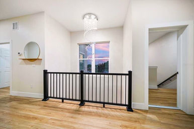 Bright upstairs landing with light wood floors, black metal railing, crystal pendant chandelier above a window showing a pink sunset, round wall mirror and open doorway to stairwell.