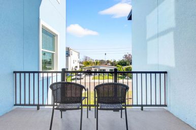 Sunlit second-floor balcony with two black wicker chairs, light-blue walls and metal railing overlooking a palm-lined suburban street under a bright blue sky.