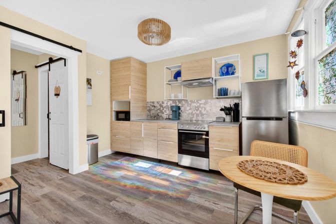 Sunlit modern kitchen with light wood cabinets, stainless steel fridge and oven, mosaic tile backsplash, open shelving with blue dishware, sliding barn door, round wood dining table with rattan chair and colorful light reflections on the wood-look floor.