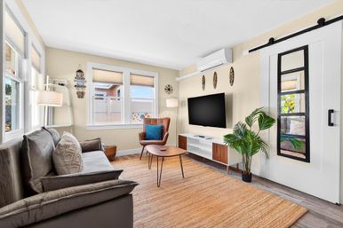 Sunny coastal-style living room with gray sofa and textured pillows, leather accent chair with blue cushion, wall-mounted TV over mid-century console, sliding barn door with window, potted palm, large windows and woven area rug on wood floors.
