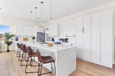 Bright open-concept modern white kitchen with a long veined island, four brown leather bar stools, hanging glass pendant lights, stainless refrigerator, tall white cabinets, hardwood floors and a potted plant.