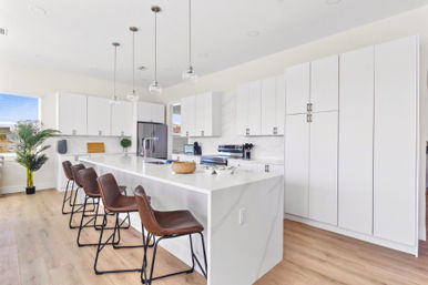 Bright open-concept modern white kitchen with a long veined island, four brown leather bar stools, hanging glass pendant lights, stainless refrigerator, tall white cabinets, hardwood floors and a potted plant.