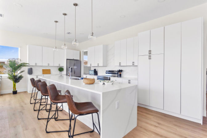 Bright open-concept modern white kitchen with a long veined island, four brown leather bar stools, hanging glass pendant lights, stainless refrigerator, tall white cabinets, hardwood floors and a potted plant.