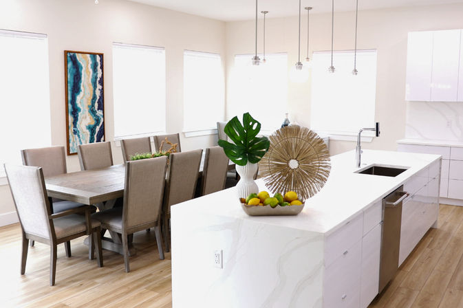 Bright modern open-plan kitchen and dining room with a white marble island and sink, hanging pendant lights, decorative sunburst sculpture, vase with monstera leaf and bowl of lemons and limes, adjacent wood dining table with eight upholstered chairs and large blue abstract wall art on light wood floors.