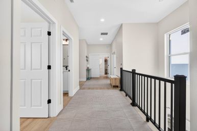 Bright second-floor hallway in a modern suburban home with beige walls, white paneled doors, carpet runner, black metal railing, small bench and recessed ceiling lights.