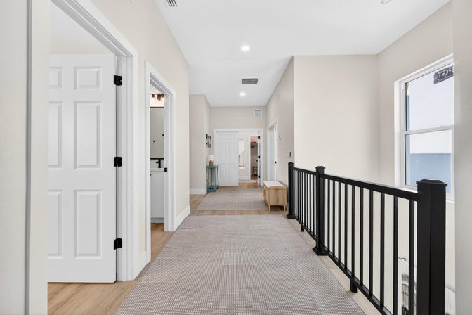 Bright second-floor hallway in a modern suburban home with beige walls, white paneled doors, carpet runner, black metal railing, small bench and recessed ceiling lights.