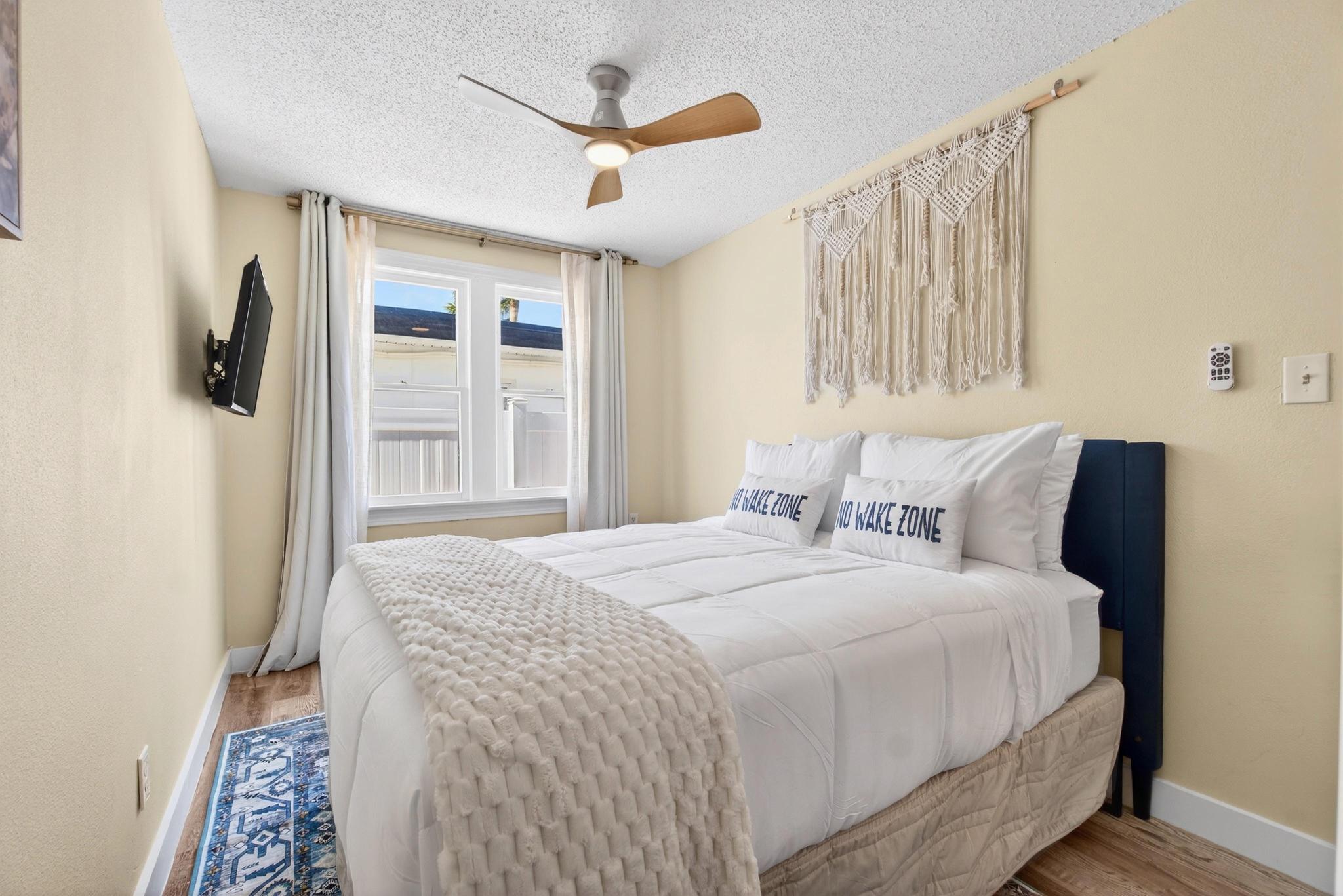 Sunlit coastal bedroom with white bedding, "NO WAKE ZONE" pillows, macramé wall hanging, wooden ceiling fan and wall-mounted TV