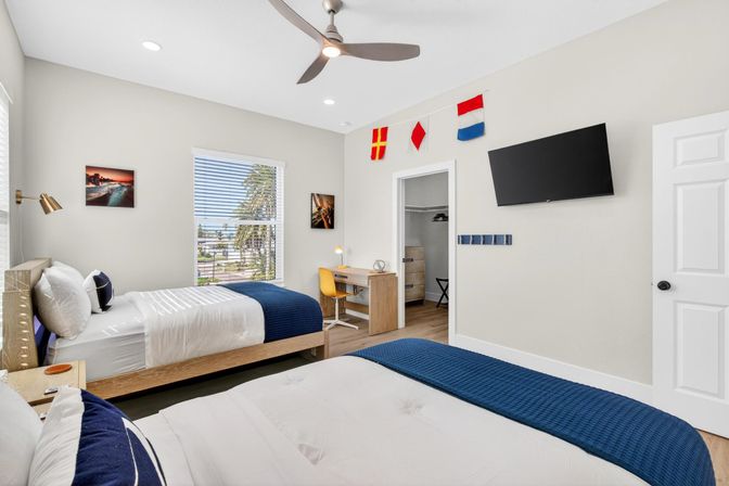 Sunny coastal-style guest bedroom with two twin beds in navy throws, wooden desk and yellow chair, wall-mounted TV, ceiling fan, and window view of palm trees and nearby street.