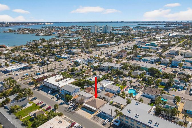 Aerial view of a sunny coastal beach town with a bay and islands in the background, showing dense rooftops, pools and streets; a red arrow marks a single-story property near the water.
