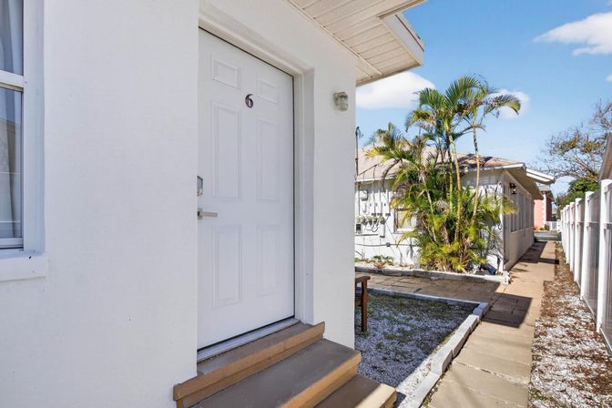 Sunny white exterior door marked 6 with steps leading to a paved walkway lined by palm trees and a white fence in a tropical residential courtyard.