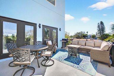 Sunny rooftop balcony patio with metal dining table and swivel chairs, wicker sectional sofa with cushions, small fire pit table, gas grill and blue patterned outdoor rug, overlooking palm trees and nearby homes.