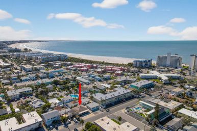 Aerial shot of a sunny beachfront neighborhood with low-rise condos, palm-lined streets and a wide sandy beach and blue ocean; a red arrow points to a single-story property near the shore, suggesting a vacation rental or seaside listing.