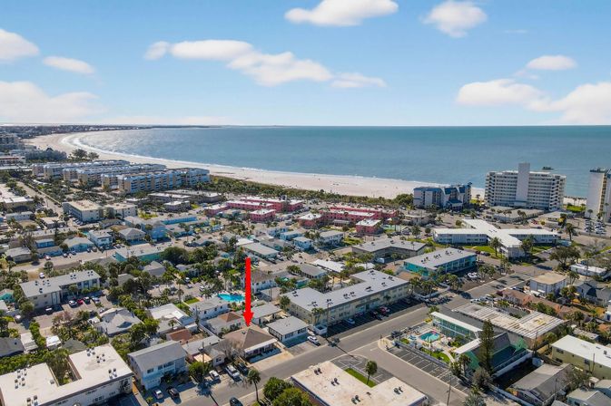 Aerial shot of a sunny beachfront neighborhood with low-rise condos, palm-lined streets and a wide sandy beach and blue ocean; a red arrow points to a single-story property near the shore, suggesting a vacation rental or seaside listing.