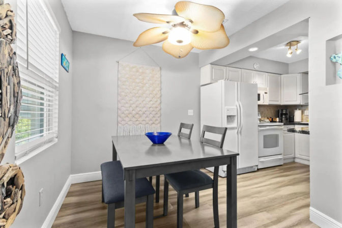 Bright open-plan dining nook with gray table and bench, blue bowl centerpiece, wood-look floors, white kitchen cabinets and a tropical leaf ceiling fan