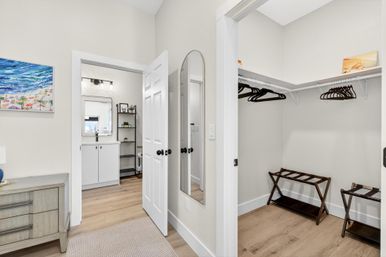 Bright modern bedroom entry with light hardwood floors, full-length mirror, open walk-in closet with wooden luggage racks and black hangers, and an en-suite bathroom vanity with coastal wall art.