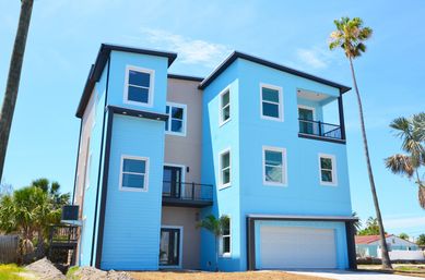 Bright blue three-story modern beach house with attached garage, balconies and palm trees under a clear sunny sky