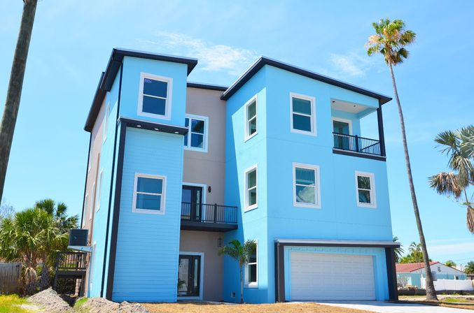 Bright blue three-story modern beach house with attached garage, balconies and palm trees under a clear sunny sky