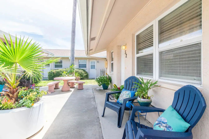 Sunny courtyard patio with blue Adirondack chairs and colorful pillows, potted tropical plants, and a round concrete picnic table beside a light-colored stucco home