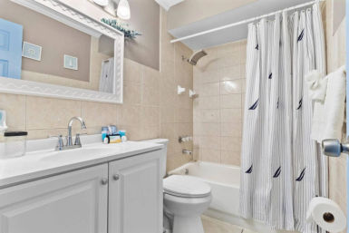 Bright coastal-inspired small bathroom with white vanity and chrome faucet, framed mirror, tiled bathtub/shower and striped shower curtain with navy sailboat motif.