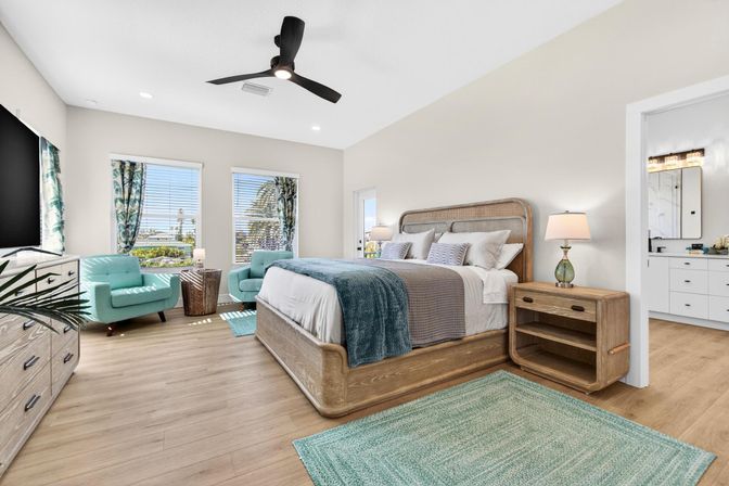 Sunny coastal master bedroom with a king bed and rattan headboard, teal throw and pillows, matching teal armchairs, wooden nightstand and dresser, ceiling fan, hardwood floors, large windows with palm-tree and water views, and a visible en-suite bathroom doorway.