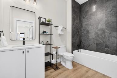 Modern bathroom with white vanity and mirror, black fixtures and open shelving, white toilet with wood-and-metal accent stand, white bathtub framed by dark gray slate tile wall, and light wood-look flooring.
