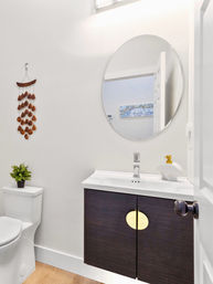 Compact modern bathroom with white walls, round mirror reflecting a hallway, dark wood floating vanity with round brass accent, white sink and faucet, toilet topped with a small potted plant, wood floor and decorative hanging wall art.