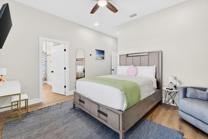 Cozy bright modern master bedroom with upholstered platform bed dressed in white linens, green throw and pink round pillow, wooden floors and blue area rug, gray armchair and bedside table with lamp and flowers, ceiling fan and wall-mounted TV; open doorway shows adjoining bathroom.