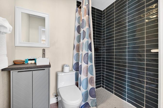 Compact modern bathroom with white toilet and gray vanity, mirror and sink, walk-in shower with dark horizontal subway tiles, hexagon floor tiles and a colorful circular-pattern shower curtain.