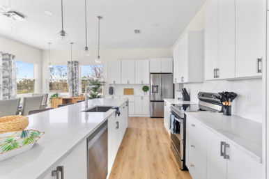 Bright open‑concept modern white kitchen featuring a large quartz island with undermount sink, pendant lights, stainless steel refrigerator and range, light wood floors and sunlit dining nook