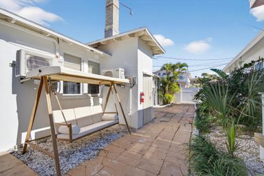 Sunny backyard patio with cushioned wooden canopy swing, stone paver walkway, tropical palms and wall-mounted air conditioners under a blue sky.