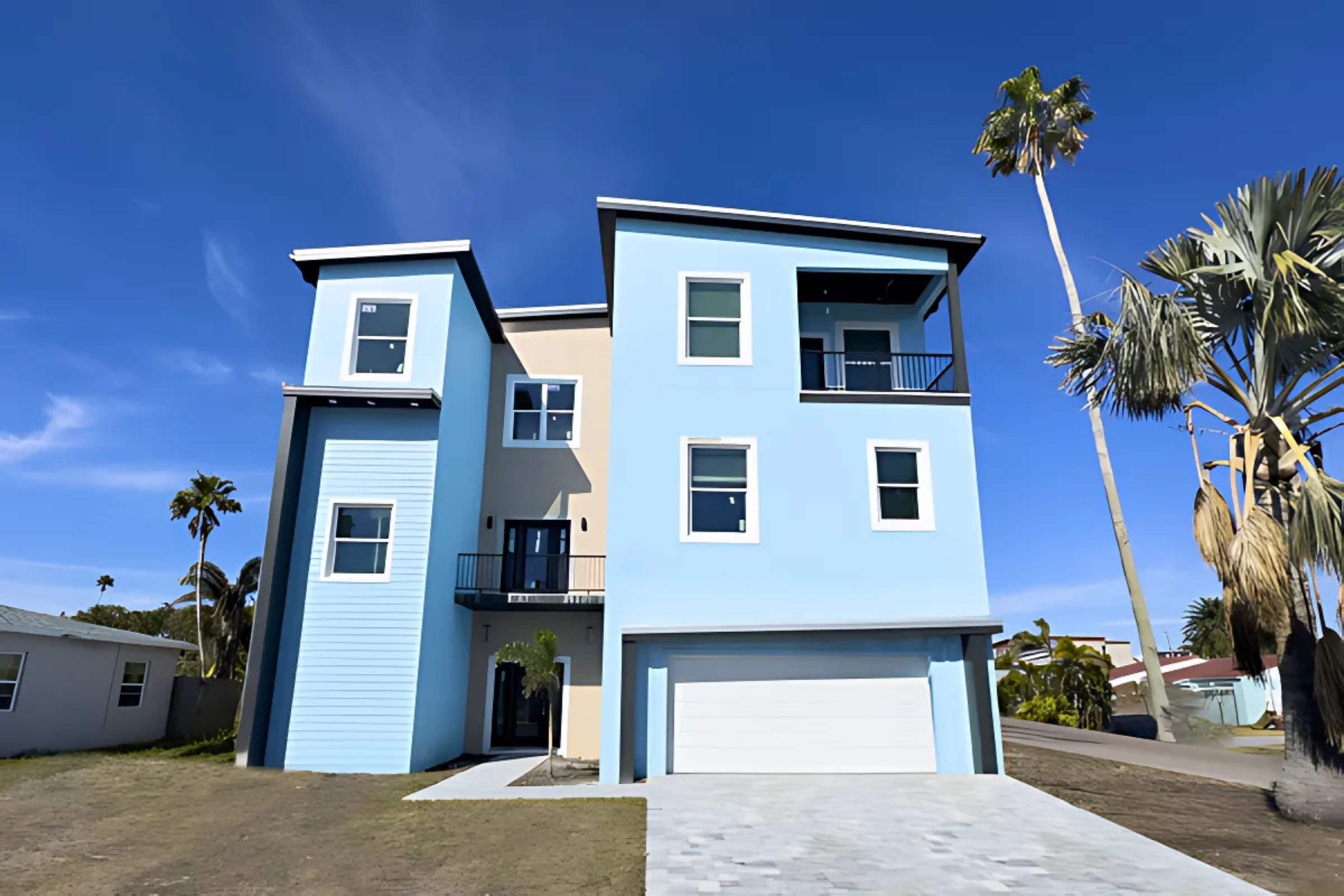 Sunny tropical three-story light-blue beach house with two-car garage, balcony and palm trees against a clear blue sky