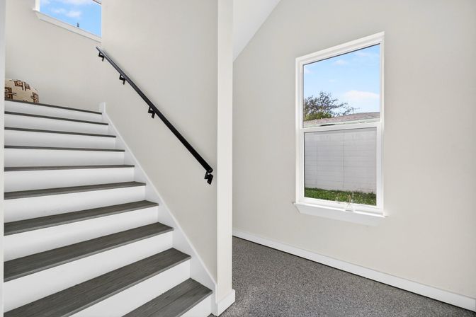 Bright modern entry with gray wood-tread staircase and black handrail, white walls, carpeted landing and window overlooking a backyard.